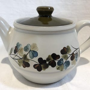 May include: A ceramic teapot with a cream-coloured body and a dark olive green lid. The teapot features a horizontal band of painted clover-like leaves in shades of blue, brown, and green wrapping around the centre of the rounded vessel.