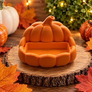 May include: A bright orange pumpkin-shaped dish with a smooth, rounded top and a hollowed-out center. The object features a textured, layered surface characteristic of 3D printing, with a prominent stem at the top and a wide, circular base.