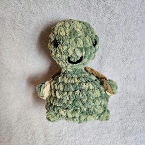 May include: A handmade plush turtle in shades of green and brown. The turtle has a smiling face with black eyes and a cream-colored patch on its flipper. The soft, textured material gives it a cozy appearance.