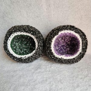 May include: Two crocheted, geode-shaped items. Each has a dark gray and white outer ring, a white inner ring, and a textured center. One center is dark green, the other is purple. The items are on a gray surface.