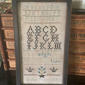 May include: Framed cross-stitch sampler with a dark wooden frame. The sampler features the alphabet in uppercase and lowercase, along with decorative elements and the text "Annie Potts 1853". The stitching is in shades of blue and brown.