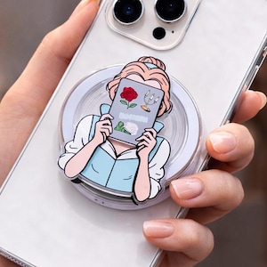 May include: A clear phone case with a pop socket featuring a cartoon illustration of a woman reading a book. The pop socket has a white border and a light blue and pink design. The book has a red rose on the cover.