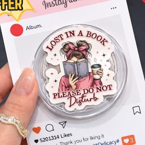 Puede incluir: Un soporte transparente para tel&eacute;fono con un gr&aacute;fico de una mujer leyendo un libro y sosteniendo una taza de caf&eacute;, con el texto "LOST IN A BOOK PLEASE DO NOT DISTURB". El soporte est&aacute; sobre un fondo rosa con tem&aacute;tica de Instagram.