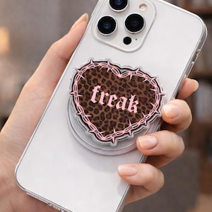 May include: A phone grip featuring a heart-shaped design with a leopard print background and the word "freak" in a gothic font. The heart is outlined with pink barbed wire.