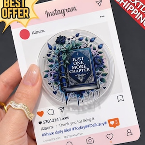 May include: A clear, round phone grip featuring a blue book with the text "JUST ONE MORE CHAPTER" surrounded by blue floral accents. The grip is set against a pink Instagram-themed background with a "BEST OFFER" sticker.