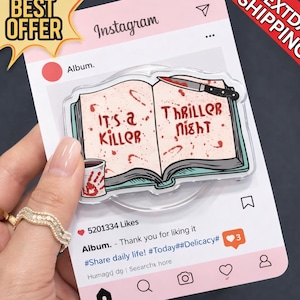 May include: A novelty Instagram-themed acrylic pin featuring a book with the text "It's a Killer Thriller Night". The book has blood splatter details and a knife. A small cup with a handprint is also shown.