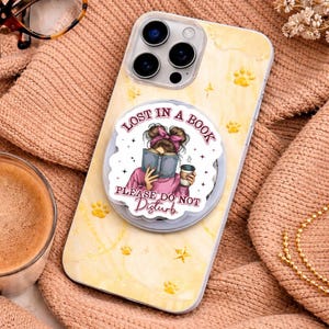 Puede incluir: Una funda de tel&eacute;fono con un agarre emergente que presenta a una mujer leyendo un libro con el texto "Lost in a Book, Please Do Not Disturb". La funda es amarilla con huellas de patas y un dise&ntilde;o de estrella. Una taza de caf&eacute; y gafas est&aacute;n en el fondo.