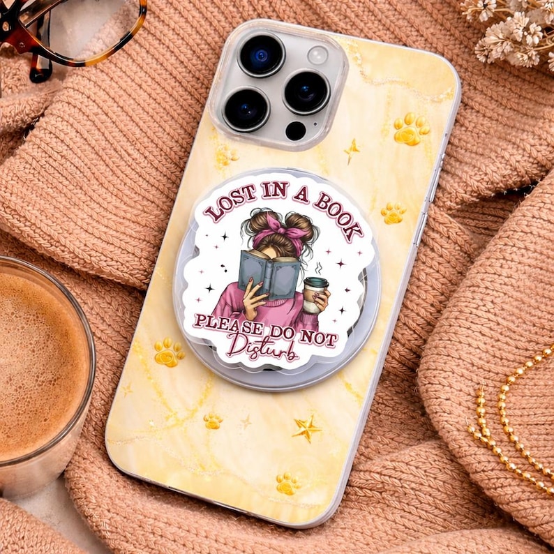 Puede incluir: Una funda de tel&eacute;fono con un agarre emergente que presenta a una mujer leyendo un libro con el texto "Lost in a Book, Please Do Not Disturb". La funda es amarilla con huellas de patas y un dise&ntilde;o de estrella. Una taza de caf&eacute; y gafas est&aacute;n en el fondo.