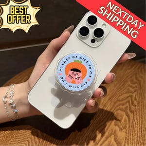 以下が含まれることがあります： 「PLEASE BE NICE TO ME OR I WILL CRY」の文字が入った、漫画のキャラクターが描かれた透明なポップアップグリップが付いた白いスマートフォン。スマートフォンにはトリプルレンズカメラが付いています。