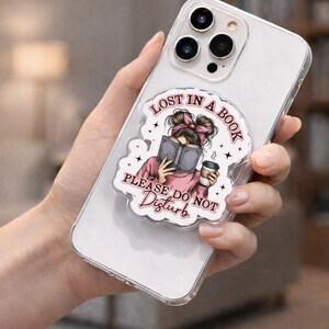 Puede incluir: Funda transparente para tel&eacute;fono con agarre desplegable. El dise&ntilde;o presenta una ilustraci&oacute;n de dibujos animados de una persona leyendo un libro, con el texto "Lost in a Book, Please Do Not Disturb". La ilustraci&oacute;n incluye un lazo rosa y una taza de caf&eacute;.
