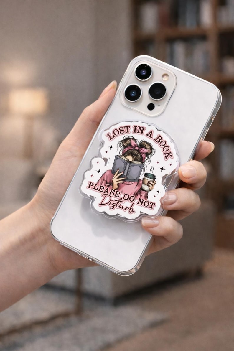 Puede incluir: Funda transparente para tel&eacute;fono con agarre desplegable. El dise&ntilde;o presenta una ilustraci&oacute;n de dibujos animados de una persona leyendo un libro, con el texto "Lost in a Book, Please Do Not Disturb". La ilustraci&oacute;n incluye un lazo rosa y una taza de caf&eacute;.