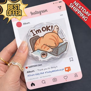 May include: A clear acrylic phone grip featuring a cartoon cat with orange stripes, a laptop, and the text "I'm OK!". The grip is designed to look like an Instagram post, with the words "Best Offer" and "Nextday Shipping" also visible.