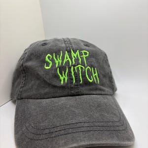 May include: A gray, distressed baseball cap with the embroidered text "SWAMP WITCH" in bright green. The hat has a curved brim and an adjustable strap at the back. The cap is made of a soft, worn fabric.