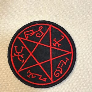 May include: A round black patch with a red pentagram design. The pentagram is outlined in red and features symbols within each point. The patch has a black border and is likely made of fabric.
