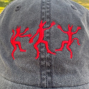 May include: A gray denim baseball cap featuring three red embroidered figures in a dancing pose. The cap has a curved brim and a structured crown. The figures are stylized with outstretched arms and legs.