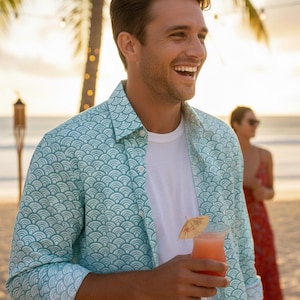 May include: A long-sleeved button-down shirt featuring a repeating teal and white fan-shaped wave pattern, worn over a plain white crew-neck t-shirt. The shirt has a classic collar, a front button placket, and cuffs that are rolled up to the elbows.