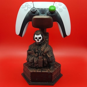 May include: A brown and black gaming controller stand featuring a bust of a character with a skull mask. The stand holds a white and black controller with green accents. The background is a solid red.