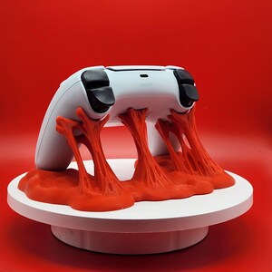 May include: A white PlayStation 5 controller rests on a white platform with a vibrant red, textured, abstract design. The controller has black thumbsticks and buttons, set against a solid red background.