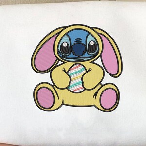 Puede incluir: Sudadera blanca con un personaje de dibujos animados con un disfraz de conejito amarillo sosteniendo un huevo de Pascua colorido. El personaje tiene piel azul, ojos grandes y detalles rosas en las orejas y patas. El diseño está centrado.