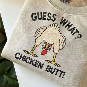 May include: A beige sweatshirt with the text "GUESS WHAT? CHICKEN BUTT!" in black. The graphic features a cartoon chicken bending over, with its rear end facing the viewer. The chicken has a red comb and yellow feet.