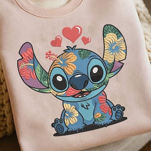 May include: A light pink sweatshirt featuring a blue cartoon character with large ears and floral patterns. The character has large black eyes and is smiling. Three red hearts float above the character's head.