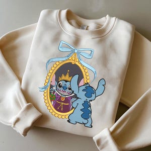 Op de afbeelding: Crèmekleurige sweatshirt met een cartoonafbeelding van Stitch die naar een spiegel kijkt met een cartoonafbeelding van Stitch met een kroon. De spiegel is goudkleurig met een blauwe strik bovenaan. Het Stitch-personage is blauw.
