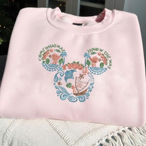 Może przedstawiać: Jasnoróżowa bluza z okrągłym dekoltem. Z przodu kolorowy haft w kształcie głowy Myszki Miki, z elementami kwiatowymi i marynistycznymi oraz napisami "COME WHAT MAY" i "I KNOW THE WAY".