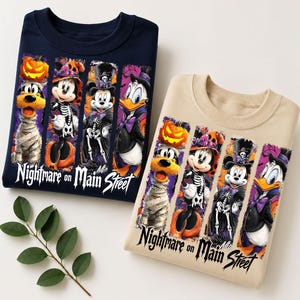 Könnte beinhalten: Ein marineblaues und ein beiges Sweatshirt mit Rundhalsausschnitt, jeweils mit einem Grafikdruck von vier Zeichentrickfiguren in Halloween-Kostümen und dem Text Nightmare on Main Street. Die Figuren sind ein Mumienhund, eine Hexe, ein Skelett und ein Vampir.