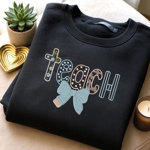 Op de afbeelding: Zwart sweatshirt met het woord "teach" geborduurd in een kleurrijk, grillig ontwerp. De letter "t" is een kruis met blauwe bloemaccenten en een lichtblauwe strik staat onderaan het woord.
