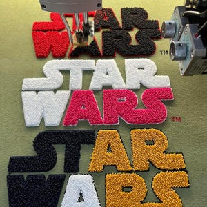 May include: An embroidery machine in action, creating the "STAR WARS" logo in multiple colours. The logo is displayed in red and black, white, pink, black and gold, and white and gold. The logo is being stitched onto a green fabric.