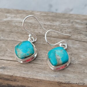 May include: A pair of turquoise and silver earrings. Each earring features a square-shaped turquoise stone set in a silver bezel. The turquoise stones have a mottled appearance with shades of blue and gray. The earrings have a hook closure.