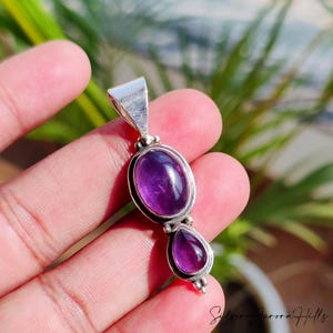May include: A silver pendant featuring two polished amethyst gemstones. The larger oval stone is above a smaller teardrop-shaped stone. The pendant has a simple, elegant design, with a bail for a chain.