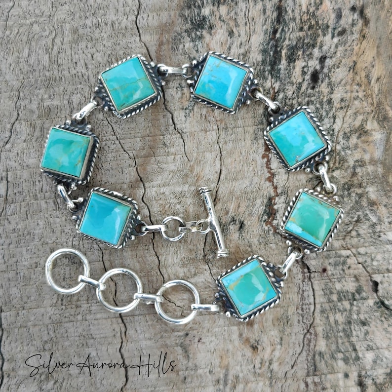 May include: A silver bracelet featuring square turquoise stones. Each stone is set in a square frame with a rope-like border. The bracelet has a toggle clasp and is displayed on a wooden surface. The text "Silver Aurora Hills" is at the bottom.