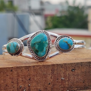 Op de afbeelding: Een zilveren manchetarmband met drie turkooizen stenen. De centrale steen heeft een onregelmatige vorm, terwijl de zijstenen rond zijn. De armband heeft een gevlochten touwontwerp rond de stenen.