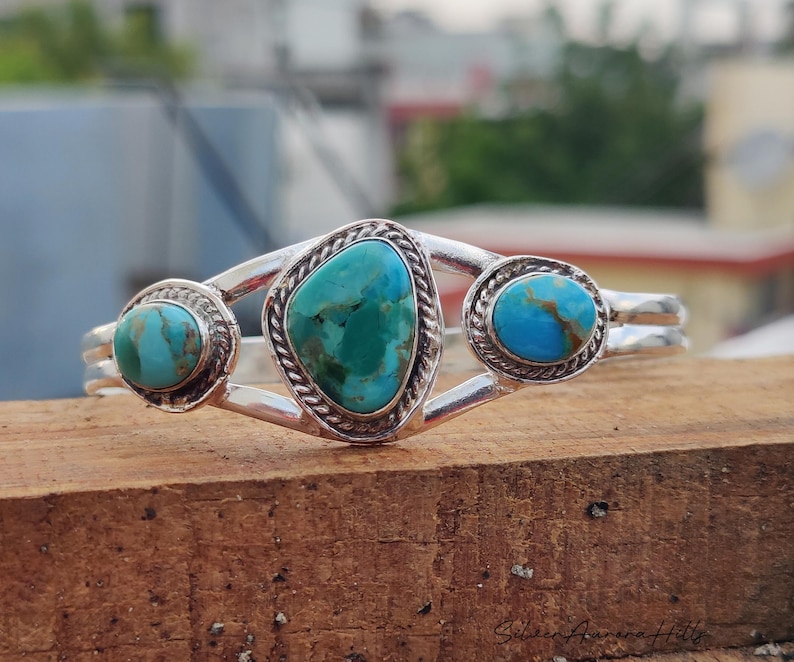 Op de afbeelding: Een zilveren manchetarmband met drie turkooizen stenen. De centrale steen heeft een onregelmatige vorm, terwijl de zijstenen rond zijn. De armband heeft een gevlochten touwontwerp rond de stenen.