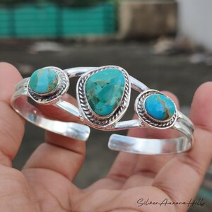 Op de afbeelding: Een zilveren manchetarmband met drie turkooizen stenen. De armband heeft een gepolijste afwerking en een decoratief ontwerp rond de turkooizen stenen. De turkooizen stenen hebben een levendige blauwgroene kleur.