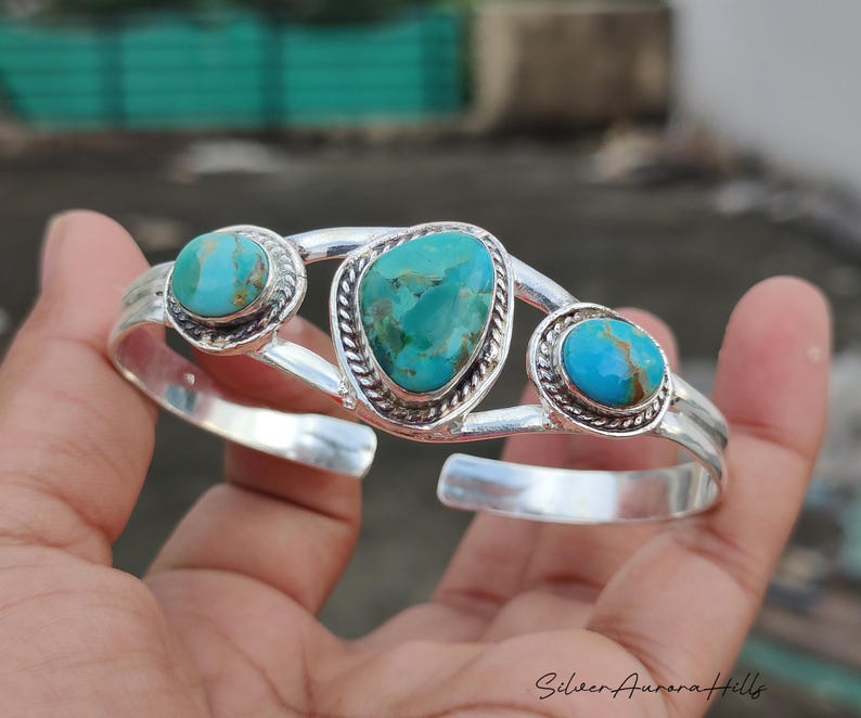 Op de afbeelding: Een zilveren manchetarmband met drie turkooizen stenen. De armband heeft een gepolijste afwerking en een decoratief ontwerp rond de turkooizen stenen. De turkooizen stenen hebben een levendige blauwgroene kleur.