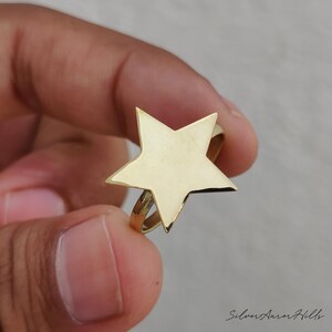 May include: A gold-toned ring featuring a large, flat, five-pointed star design centered on a thin, circular band. The star has smooth, polished surfaces and sharp, defined points. The text Silver Aurora Hills is visible in the bottom right corner.