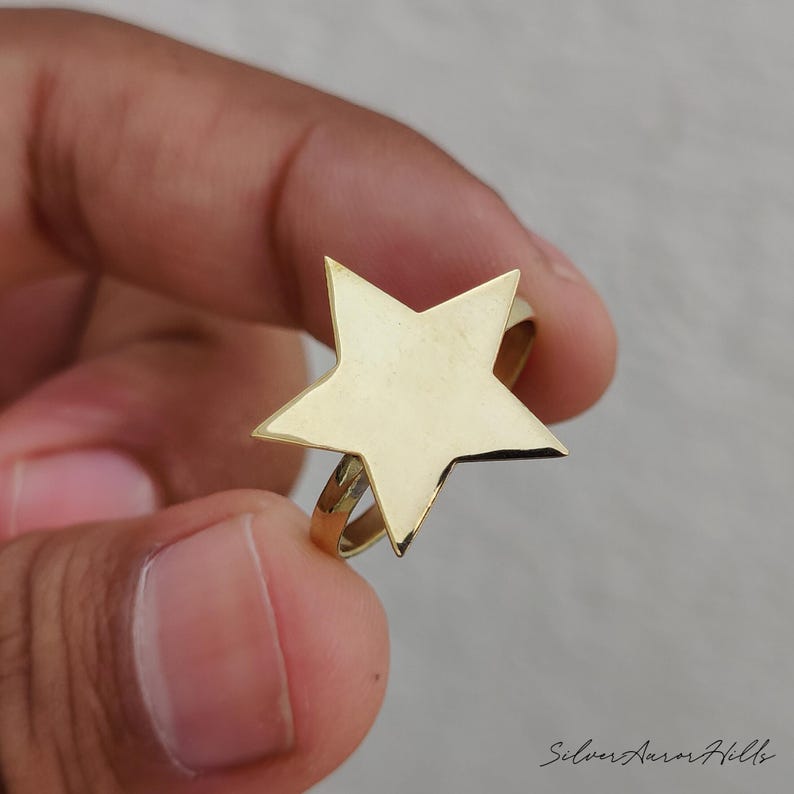May include: A gold-toned ring featuring a large, flat, five-pointed star design centered on a thin, circular band. The star has smooth, polished surfaces and sharp, defined points. The text Silver Aurora Hills is visible in the bottom right corner.