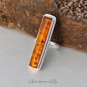 May include: A silver ring featuring a long, rectangular amber stone. The amber is a rich orange hue with internal markings. The ring is set against a neutral backdrop, with the text "Silver Aurora Hills" at the base.