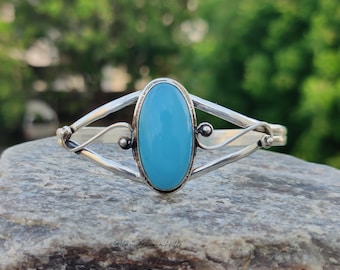 May include: A silver bracelet with a large, oval, light blue stone at the center. The bracelet has a delicate, open design with curved silver accents and small silver beads. The bracelet is displayed on a gray stone surface against a blurred green background.