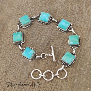 May include: A silver bracelet featuring square turquoise stones. Each stone is set in a square frame with a rope-like border. The bracelet has a toggle clasp and is displayed on a wooden surface.
