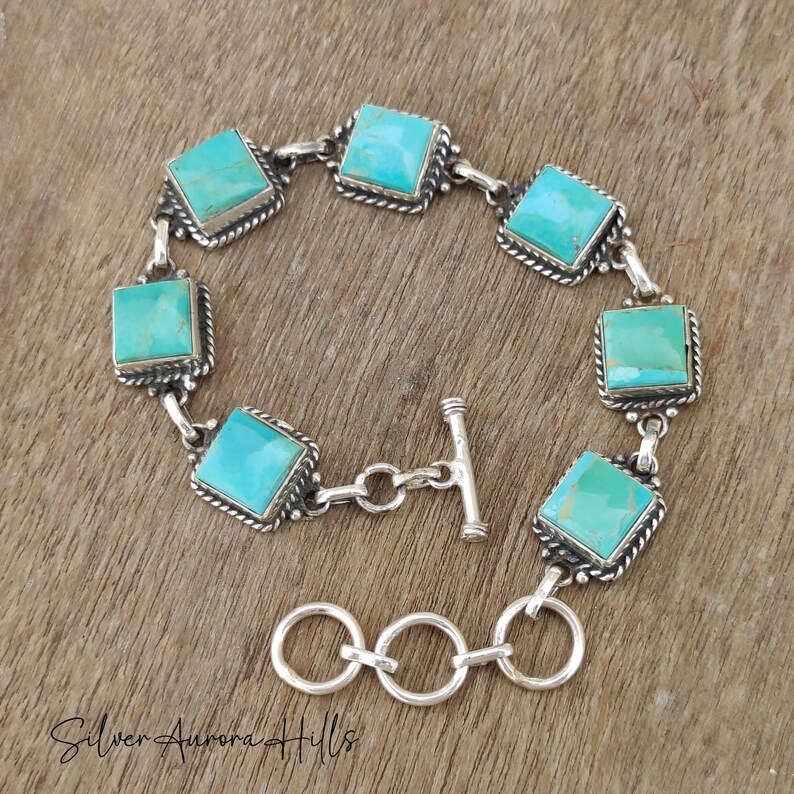 May include: A silver bracelet featuring square turquoise stones. Each stone is set in a square frame with a rope-like border. The bracelet has a toggle clasp and is displayed on a wooden surface.