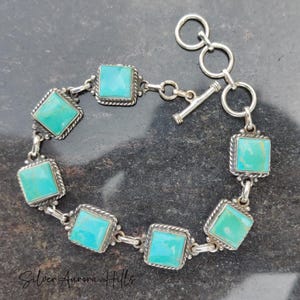 May include: A silver bracelet featuring square turquoise stones. Each stone is set in a square frame with a twisted rope design. The bracelet has a toggle clasp and is displayed on a dark surface. The bracelet is a piece of jewelry.