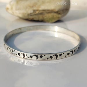 May include: A silver bangle bracelet with a celestial design of crescent moons and stars. The bracelet is circular and has a polished finish. The design is etched around the entire circumference. The background includes a light-coloured stone.