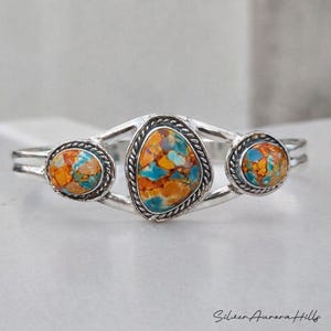 Op de afbeelding: Zilveren armband met drie kleurrijke stenen in oranje, blauw en bruin. De centrale steen is ovaal, de zijstenen zijn rond. De armband heeft een decoratieve, touwachtige rand. Een sieraad.