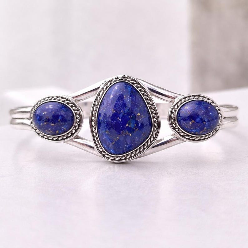 Op de afbeelding: Zilveren manchetarmband met drie diepblauwe lapis lazuli stenen. De centrale steen heeft een onregelmatige vorm, geflankeerd door twee ovale stenen. Elke steen is gezet in een zilveren bezel met een touwachtige rand.