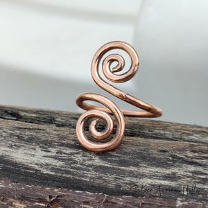 May include: A copper ring with a double spiral design. The ring features two swirling spirals connected by a curved band. The ring is displayed on a weathered wooden surface.