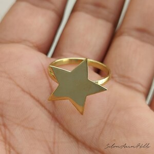 May include: A gold-toned ring featuring a flat, five-pointed star design centered on a thin, circular band. The star has a smooth, reflective surface and sharp, defined points. The ring is presented in a clear, focused view with a polished finish.