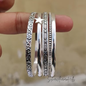 May include: A collection of silver-toned bangles, each with unique designs. One bangle features a floral pattern, another a star, and others have intricate details. The bangles are stacked together, showcasing their varied textures and styles. The image is set against a blurred background.